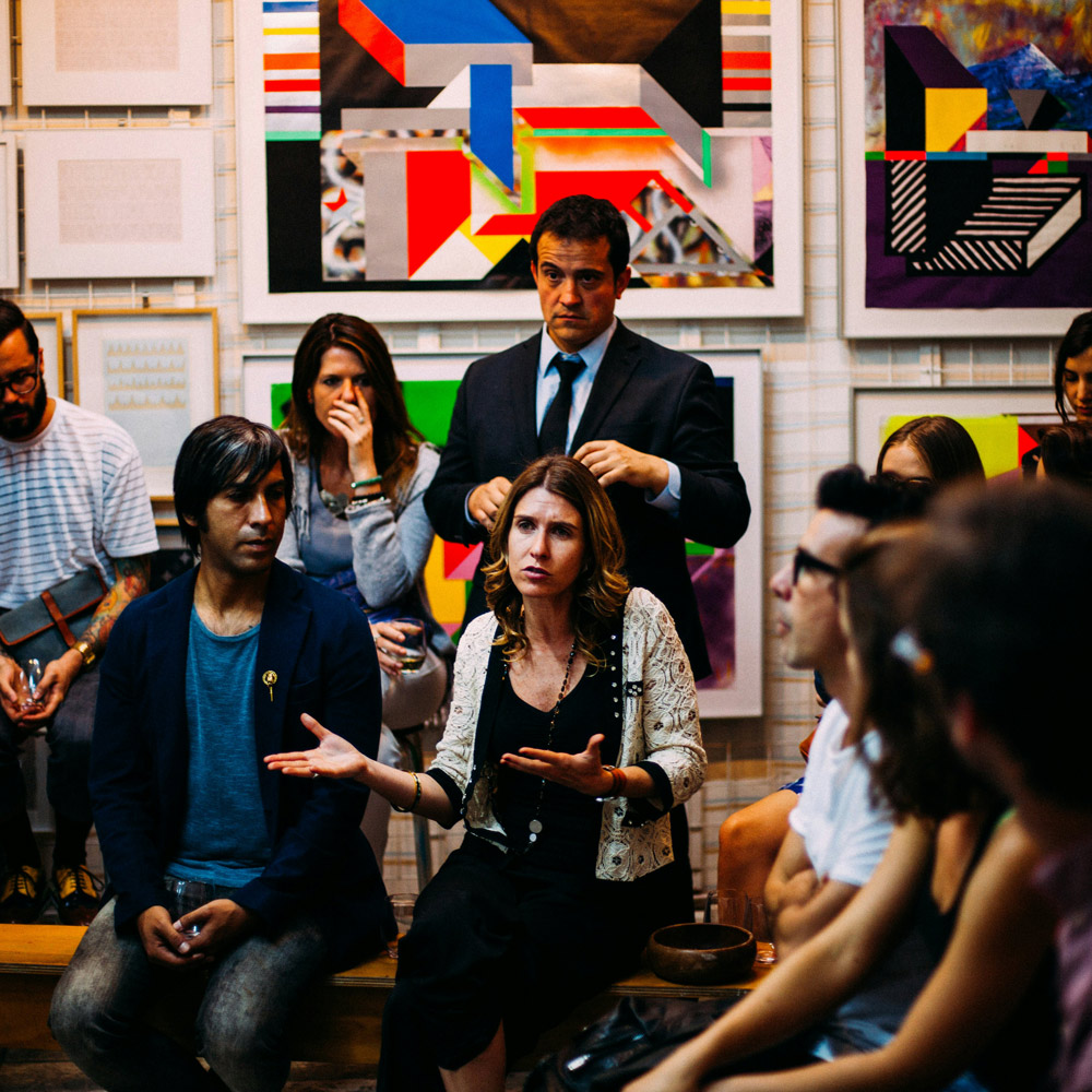 A group of people sitting in an art gallery; one person is talking and gesturing with her hands.