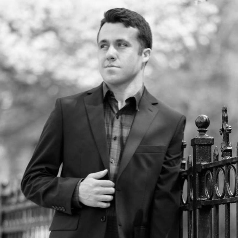 A man in a suit. He leans against a wrought-iron fence.